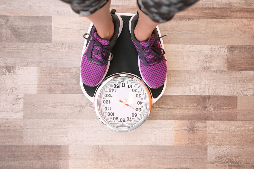 Woman measuring her weight using scales on floor, top view Person in purple sneakers standing on a mechanical scale, displaying a weight with wooden flooring in the background.